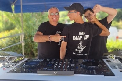 Trois personnes posent devant une table de mixage sous un chapiteau, avec une banderole ’DJ GÉI ANIMATION’.