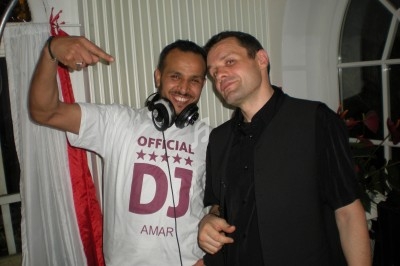 Deux hommes posent devant une table de DJ, l'un portant un casque audio et un t-shirt avec l'inscription ’Official DJ’.