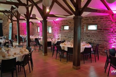 Salle de réception avec tables rondes dressées, poutres apparentes et éclairage rose sur les murs en pierre.