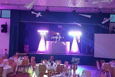 Salle de réception décorée, tables dressées avec chaises jaunes, éclairage coloré et espace DJ en arrière-plan.