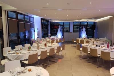 Salle de réception lumineuse avec tables rondes dressées, chaises modernes et éclairage d'ambiance.