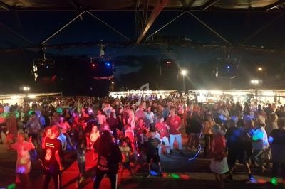 Foule dansant sous un chapiteau éclairé par des lumières colorées, avec des stands en arrière-plan.