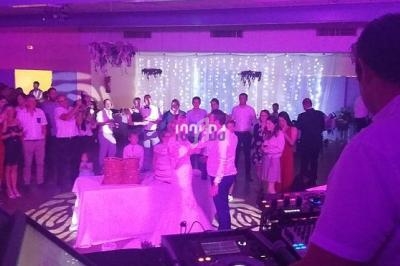 Un couple danse au centre d'une salle éclairée, entouré d'invités debout, avec un DJ au premier plan.