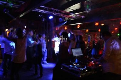 Des personnes dansent dans une ambiance festive avec un DJ en arrière-plan dans un club éclairé par des lumières colorées.