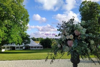 Un grand bouquet de fleurs devant une pelouse avec une tente blanche et des arbres sous un ciel partiellement nuageux.
