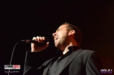 Un chanteur en costume noir interprète une chanson sur scène, tenant un micro dans une main.