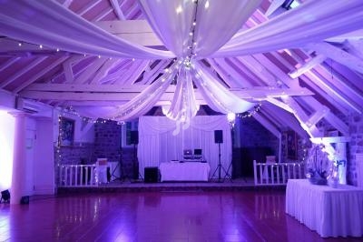 Salle de réception avec poutres apparentes, décorée de drapés blancs et guirlandes lumineuses, éclairage violet.