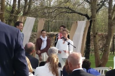 Une femme lit un discours au micro lors d'une cérémonie en extérieur, entourée de quelques personnes et d'invités assis.