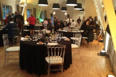 Salle de réception avec tables rondes dressées, invités debout et assis, éclairage chaleureux et décor élégant.