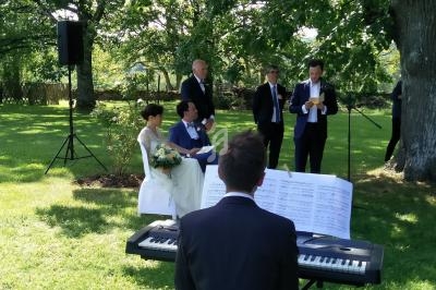 Un couple assis lors d'une cérémonie en plein air, un homme joue du piano et un autre lit un discours au micro.