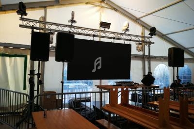 Écran noir suspendu dans une structure métallique sous une tente, entouré de haut-parleurs et de tables en bois.