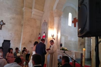 Un couple échange ses vœux devant un prêtre dans une église, entouré d'invités assis et de décorations sobres.