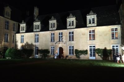 Façade d'un bâtiment historique en pierre éclairé la nuit, avec des fenêtres à lucarnes et un jardin au premier plan.