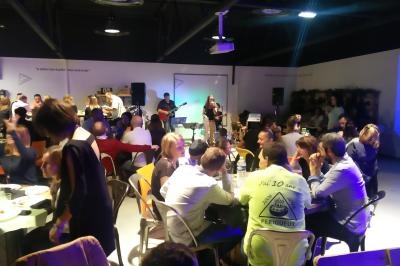 Groupe de musique jouant sur scène dans une salle animée, avec des convives attablés et discutant.