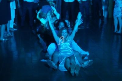 Des personnes assises en file sur le sol, bras levés, participant à une animation festive dans une salle éclairée en bleu.