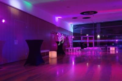 Salle moderne éclairée par des lumières violettes et roses, avec tables hautes et basses disposées pour un événement.