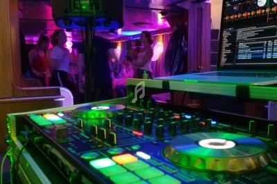 Console de mixage éclairée au premier plan, avec des invités dansant et discutant dans une ambiance lumineuse festive.