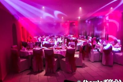 Salle de réception décorée avec des tables rondes, nappes blanches, chaises ornées de nœuds rouges et éclairage rose.