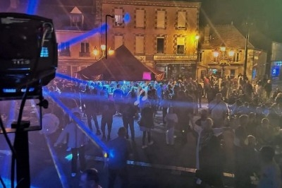 Foule rassemblée de nuit sur une place illuminée, avec des jeux de lumière et des bâtiments en arrière-plan.