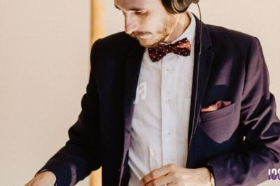 Un DJ en costume et nœud papillon manipule des équipements audio dans un environnement intérieur.