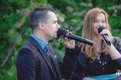 Un homme et une femme chantent en duo avec des micros, debout devant un fond de verdure.