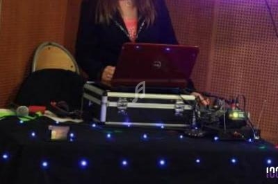 Femme debout derrière une table équipée d'un ordinateur portable et de matériel audio sous une lumière tamisée.