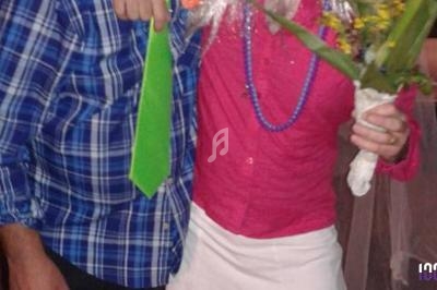 Un homme en chemise à carreaux et cravate verte pose à côté d'une personne en tenue colorée tenant un bouquet de fleurs.