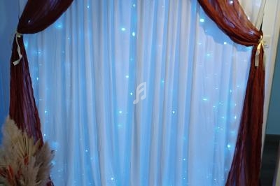 Rideau blanc illuminé de guirlandes LED bleues, encadré de voilages marron noués, avec un vase de pampas à gauche.