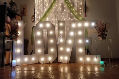 Lettres lumineuses formant le mot ’LOVE’ devant un rideau éclairé, décorées de plantes et de bougies colorées.