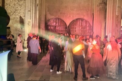 Personnes dansant dans une salle en pierre avec deux grandes portes en bois en arrière-plan, éclairées par des lumières…