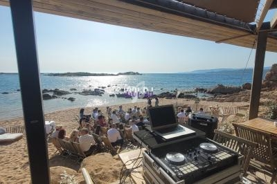 Poste de DJ en plein air sur une plage, avec des invités assis face à la mer sous un ciel ensoleillé.