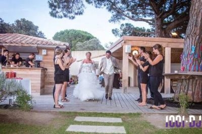 Un couple de mariés souriants entre sur une terrasse en bois, applaudi par des invités debout.
