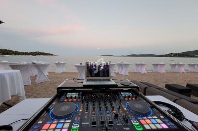Console de DJ sur une plage au coucher du soleil, avec des tables hautes et une vue sur la mer en arrière-plan.