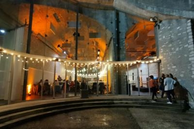 Façade d'un bâtiment en pierre avec arches illuminées et guirlandes lumineuses, des personnes discutent à l'entrée.