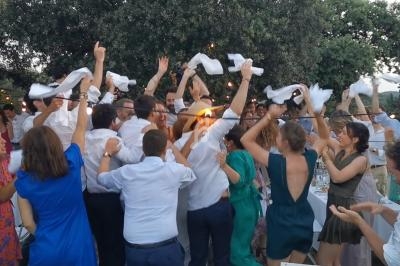 Groupe de personnes dansant et agitant des serviettes blanches en extérieur, sous des arbres et des guirlandes lumineuses.