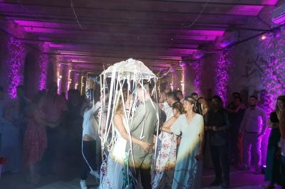 Un couple danse sous une décoration lumineuse en forme de parapluie, entouré d'invités dans une salle éclairée en violet.