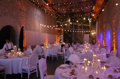 Salle de réception décorée avec des guirlandes lumineuses, tables rondes dressées et chaises blanches élégantes.