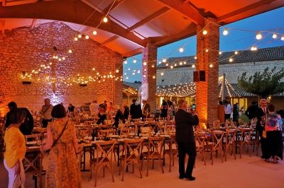 Salle en plein air décorée de guirlandes lumineuses, avec des tables dressées et des invités debout en soirée.