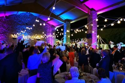 Des convives agitent des serviettes lors d'une fête de soirée sous une structure éclairée par des guirlandes lumineuses.