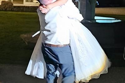 Un homme en costume soulève une femme en robe de mariée dans une cour extérieure éclairée.