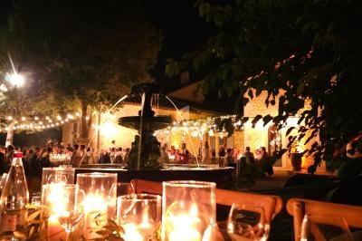 Table dressée avec bougies et verres, en extérieur, éclairée par des guirlandes lumineuses et entourée de végétation.