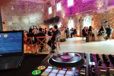 Salle en pierre avec des invités assis à des tables rondes, vue depuis une table de mixage de DJ au premier plan.