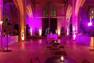 Salle voûtée éclairée de lumières violettes, avec tables et chaises disposées pour un événement.