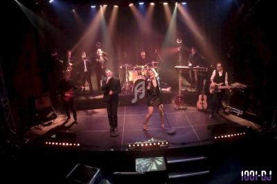 Groupe musical en concert sur scène avec chanteurs, musiciens et éclairage coloré.