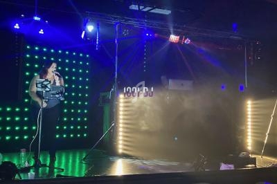 Une femme chante sur scène avec un micro, éclairée par des lumières colorées, devant un DJ et des équipements.