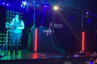 Une femme chante sur scène avec un micro, accompagnée d'un DJ en arrière-plan sous des lumières colorées.