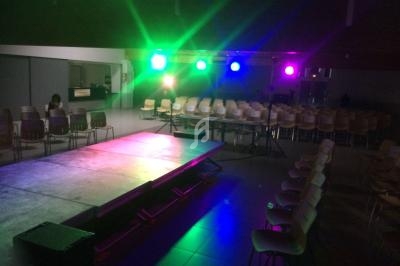 Scène éclairée par des projecteurs colorés dans une salle avec des rangées de chaises vides.