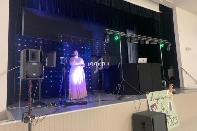 Une chanteuse en robe rose se produit sur scène avec des lumières colorées et un équipement de sonorisation.