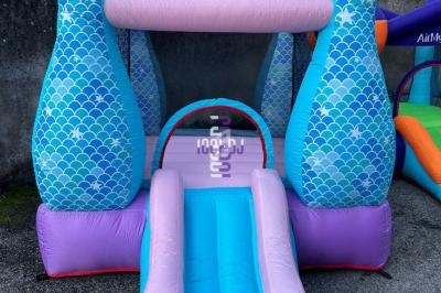 Structure gonflable bleue et rose en forme de queue de sirène avec un toboggan, installée sur un sol extérieur.