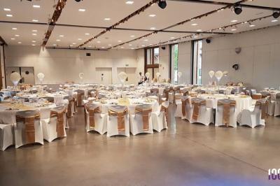 Salle de réception décorée avec des tables rondes, nappes blanches et chaises ornées de rubans dorés.